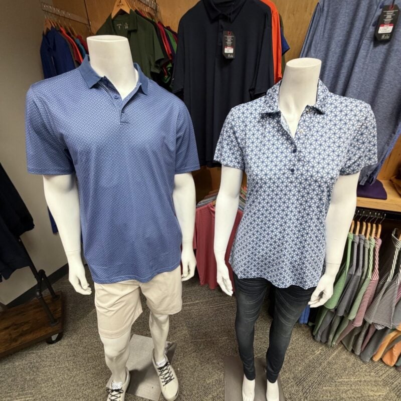 Photo from an AGM Regional Event at Antigua Apparel, showing two mannequins wearing men's and women's blue patterned golf polos, paired with khaki shorts and dark pants. Additional Antigua apparel is displayed on racks in the background.