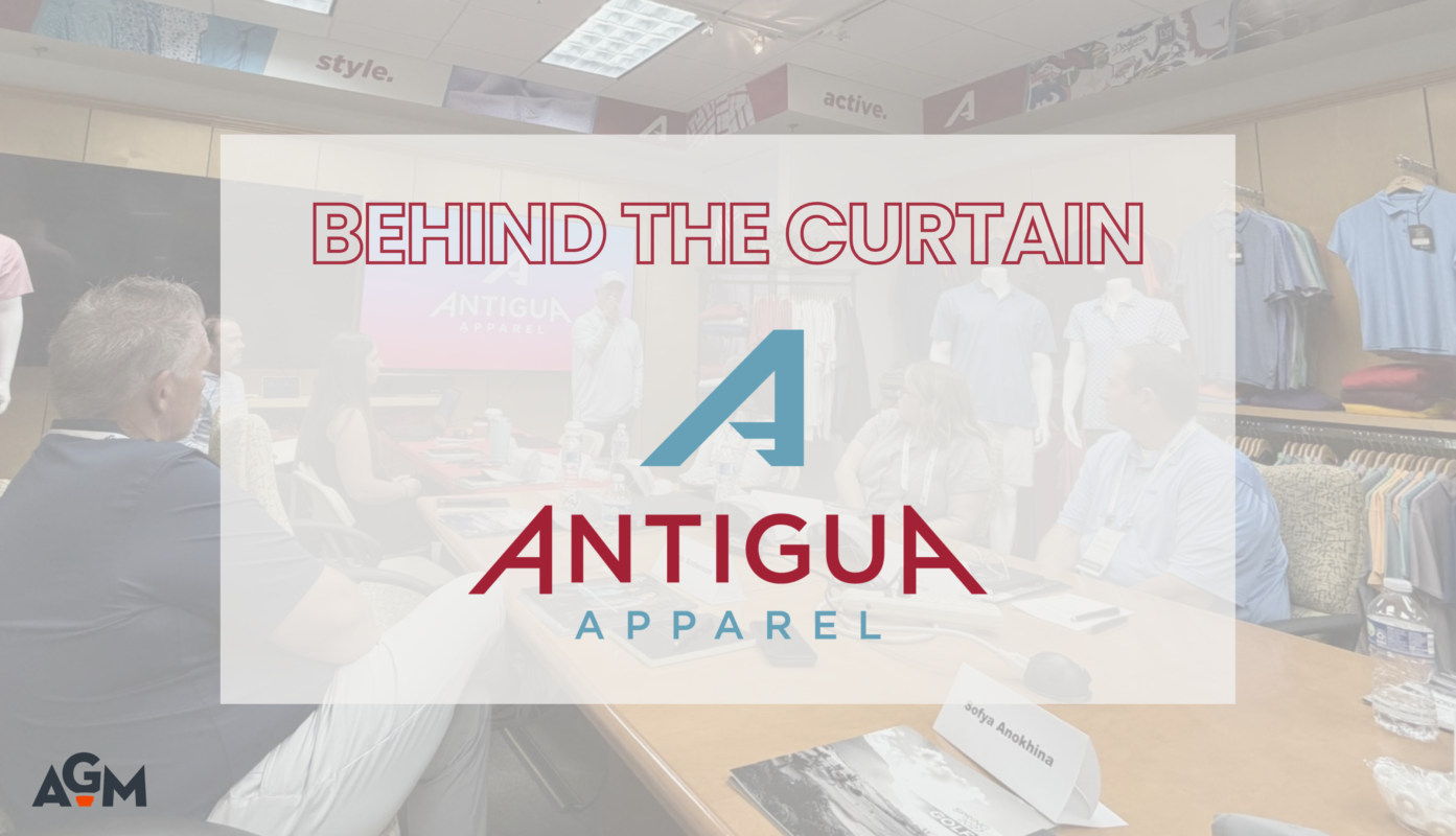 Featured graphic for the article “Behind the Curtain at Antigua,” featuring the Antigua Apparel logo over a faded photograph of AGM members attending a presentation at Antigua's facility. The AGM logo appears in the bottom left corner.