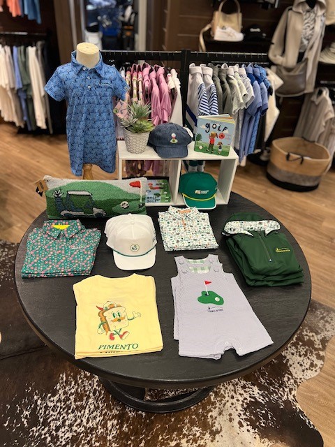 Photograph of a children's golf apparel display featuring colorful patterned polos, rompers, shorts, hats, and golf-themed accessories arranged on a round table. A small mannequin wears a blue printed romper, with additional pink, blue, and neutral-colored children's clothing hanging on racks behind the display.