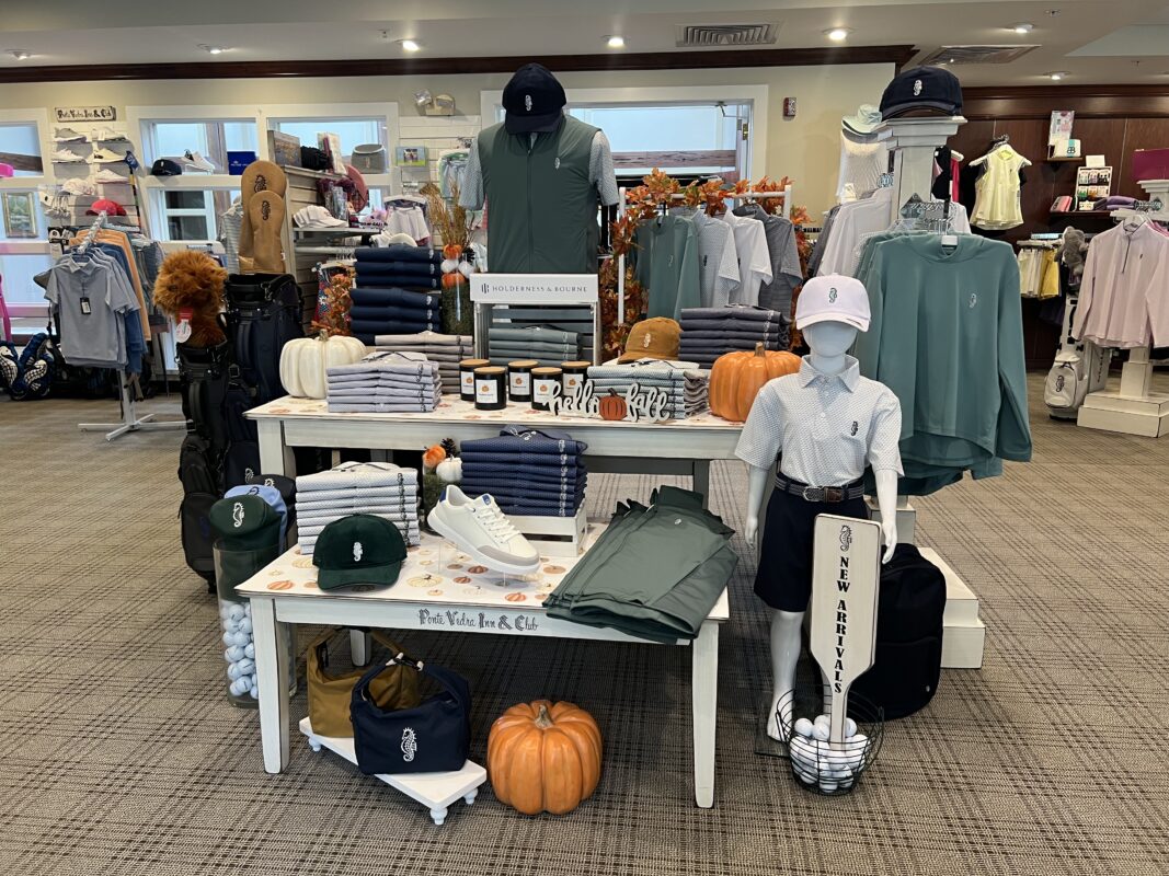 Photograph of a fall-themed golf shop display at Ponte Vedra Inn & Club in Ponte Vedra Beach, Florida, featuring neatly folded Holderness & Bourne apparel, hats, golf shoes, and accessories in navy, green, and white. Pumpkins, autumn foliage, and a “New Arrivals” sign decorate the display, with mannequins showcasing golf apparel.