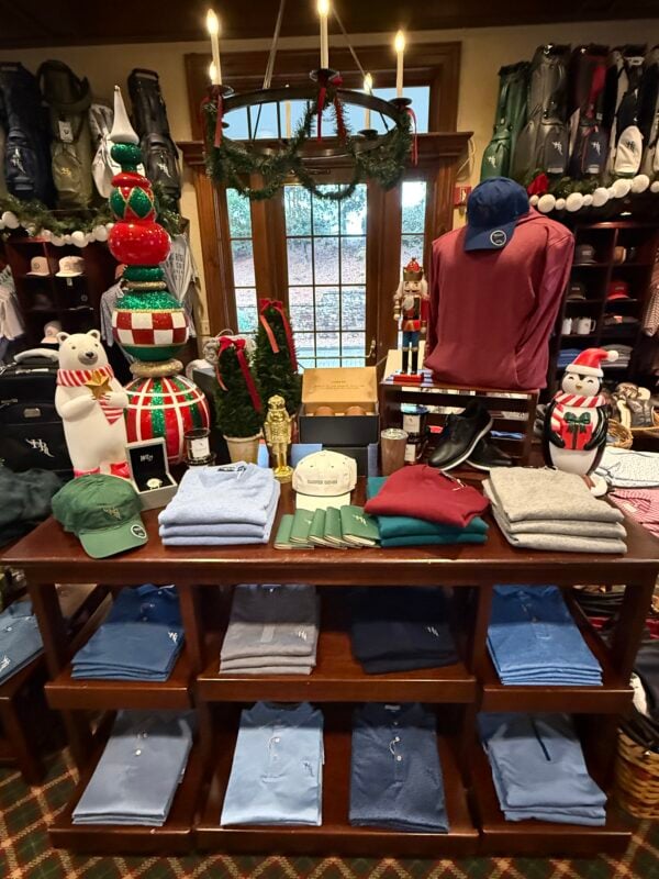 Holiday golf shop display at Hawks Ridge Golf Club featuring neatly folded apparel, golf hats, shoes, and accessories arranged on a wooden table with festive nutcrackers, a polar bear, a penguin, colorful ornaments, and a garland-wrapped chandelier.