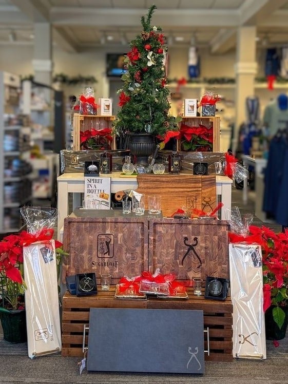 Holiday golf shop display at TPC Sugarloaf featuring a decorated Christmas tree, red poinsettias, branded wooden boxes, drinkware, and gift items arranged on tiered wooden crates with red ribbon accents.